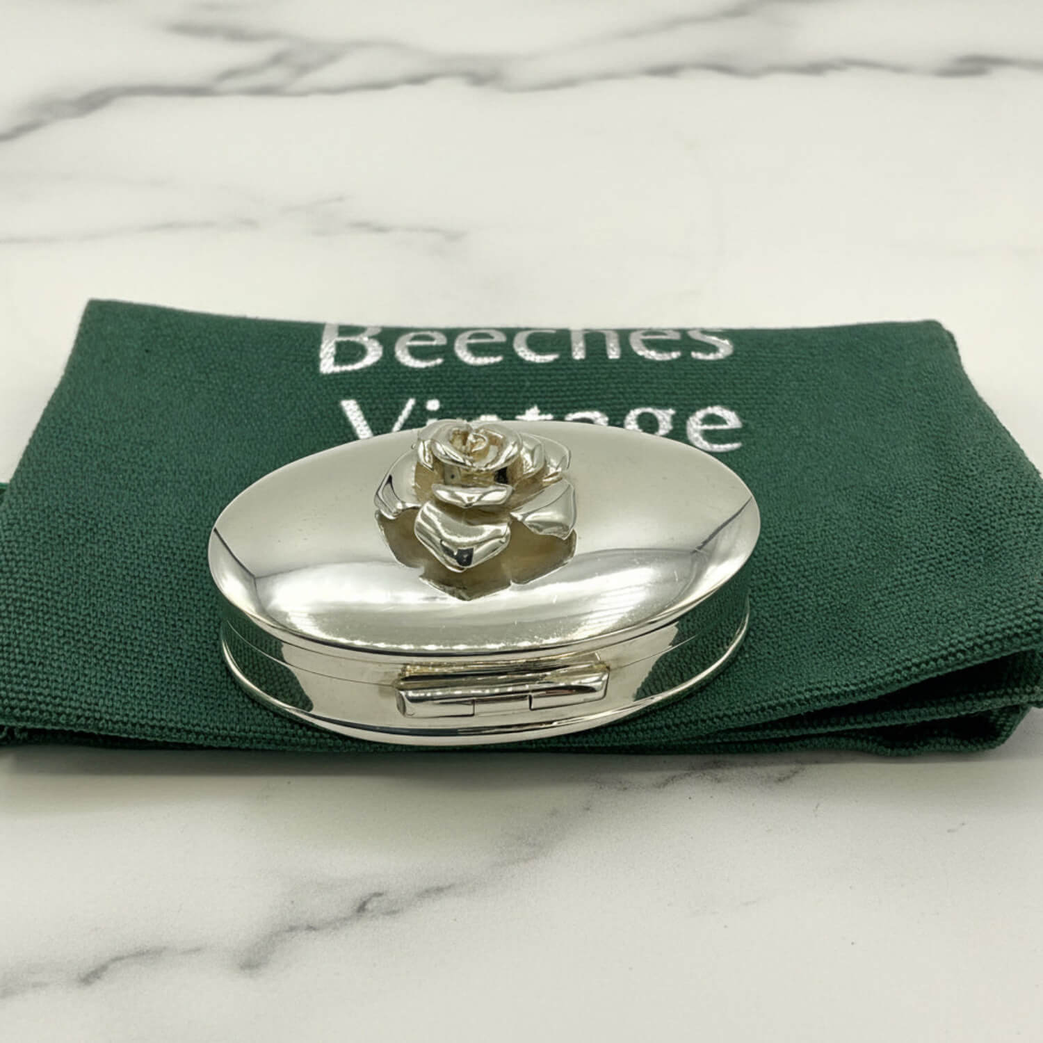 Silver oval pill box with rose design on a green cloth bag with 'Beeches Vintage' branding.