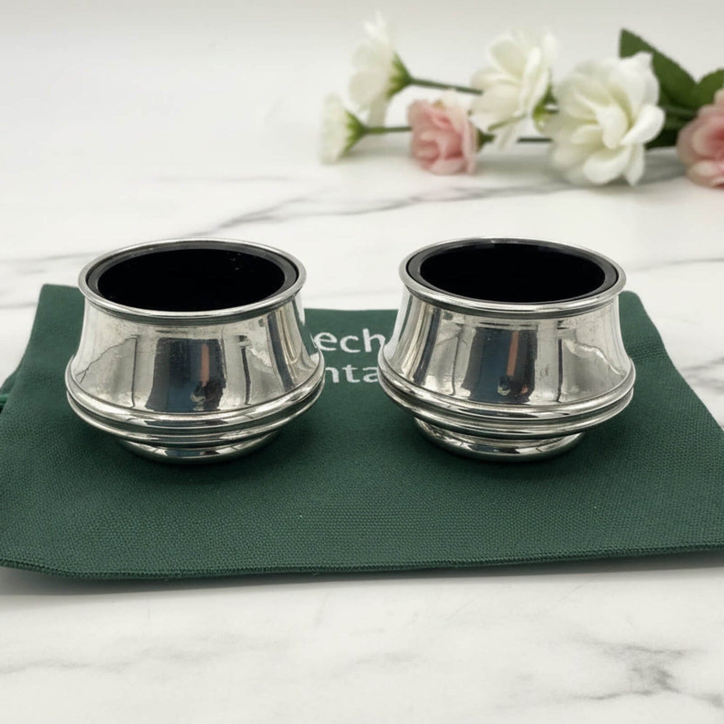 Two silver salt shakers with blue liners on a green cloth with flowers in the background.