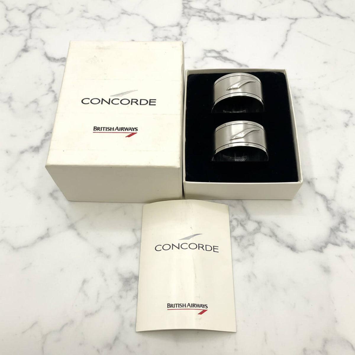 A pair of silver metal Concorde British Airways napkin rings in a black-lined presentation box, shown with the branded outer box and folded paper insert on a white background.