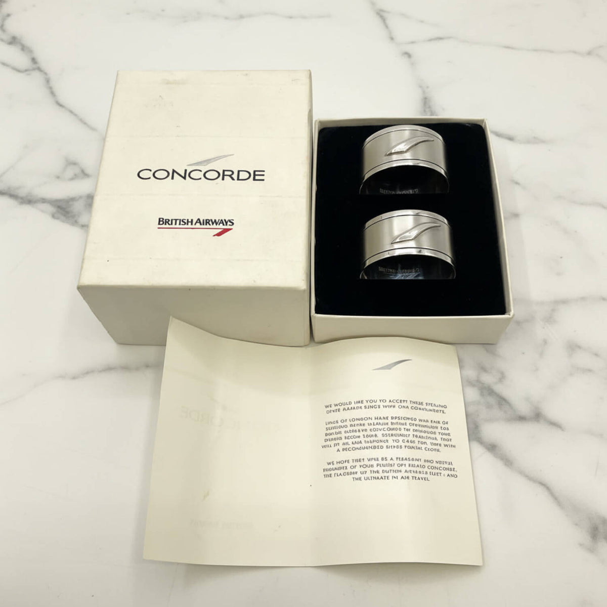 A pair of silver Concorde British Airways napkin rings presented in a black box next to their cream-colored branded packaging box and a collector's booklet.