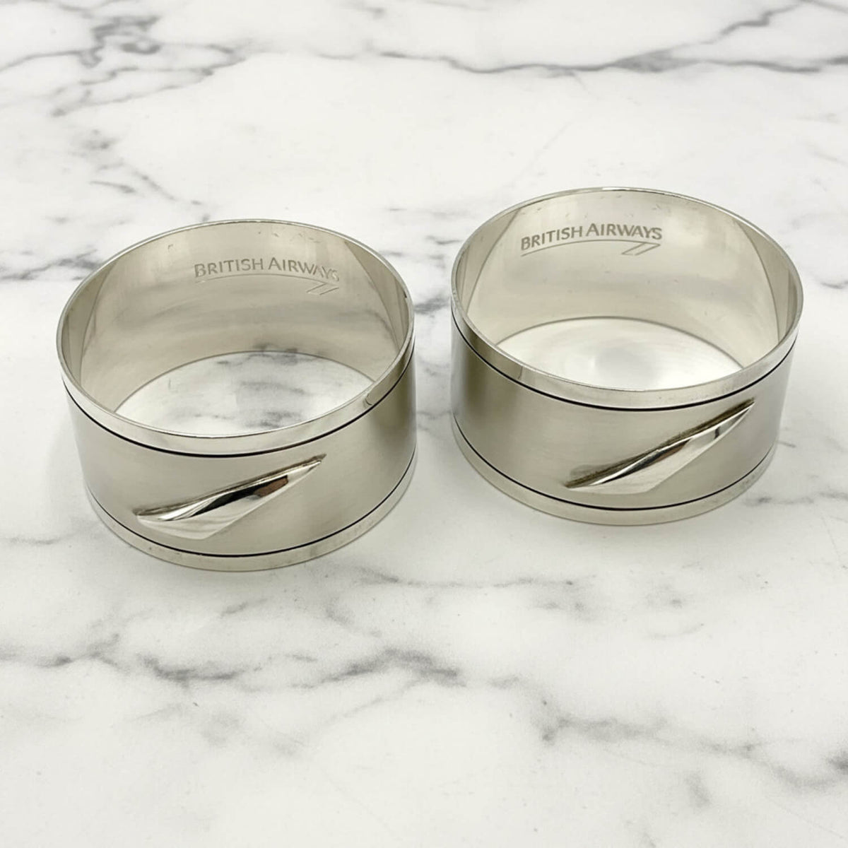 A pair of vintage silver British Airways napkins rings with a sleek diagonal cutout design and the brand name engraved inside the rim, set against a plain white background.
