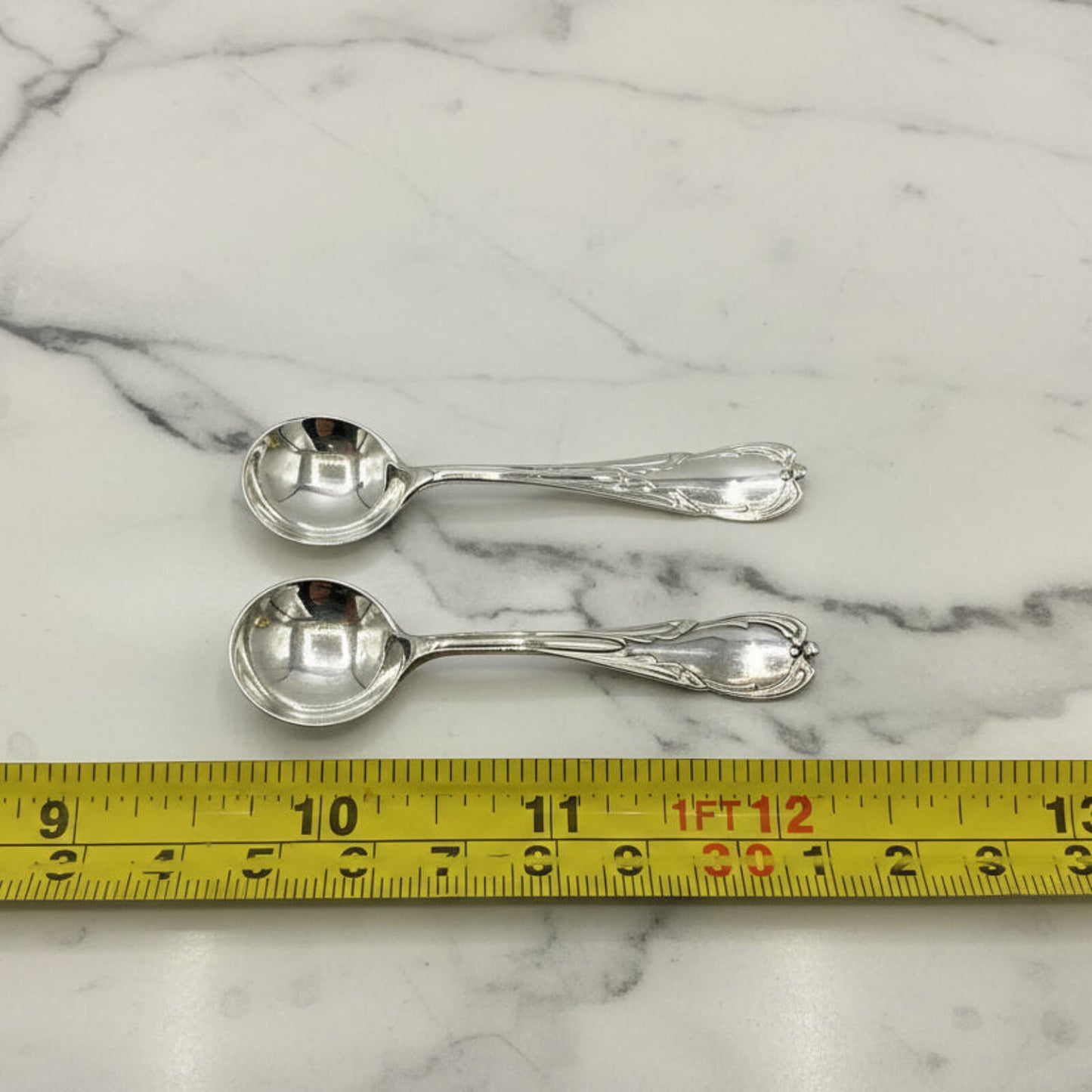 Two silver spoons on a marble surface with a tape measure for scale.