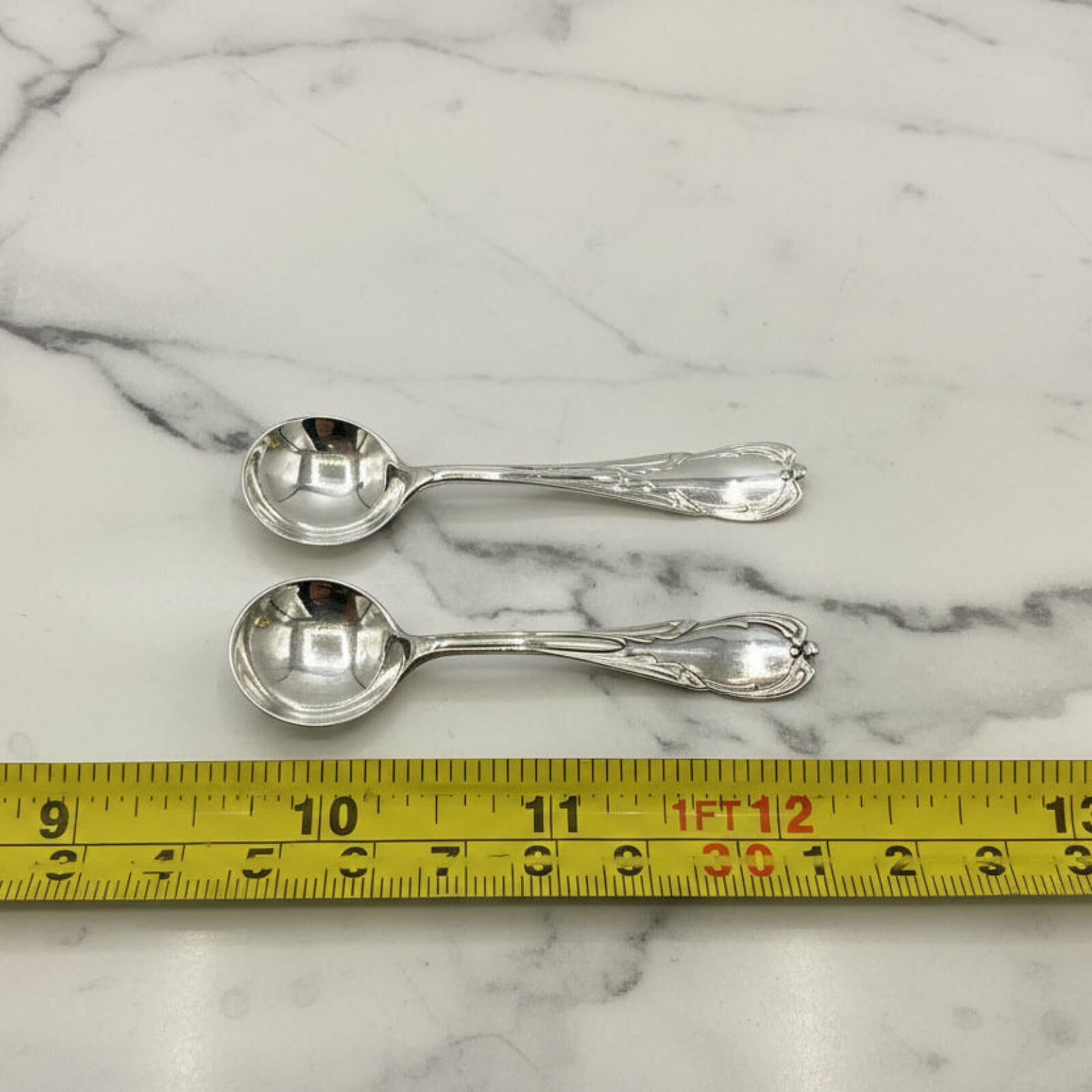 Two silver spoons on a marble surface with a tape measure for scale.