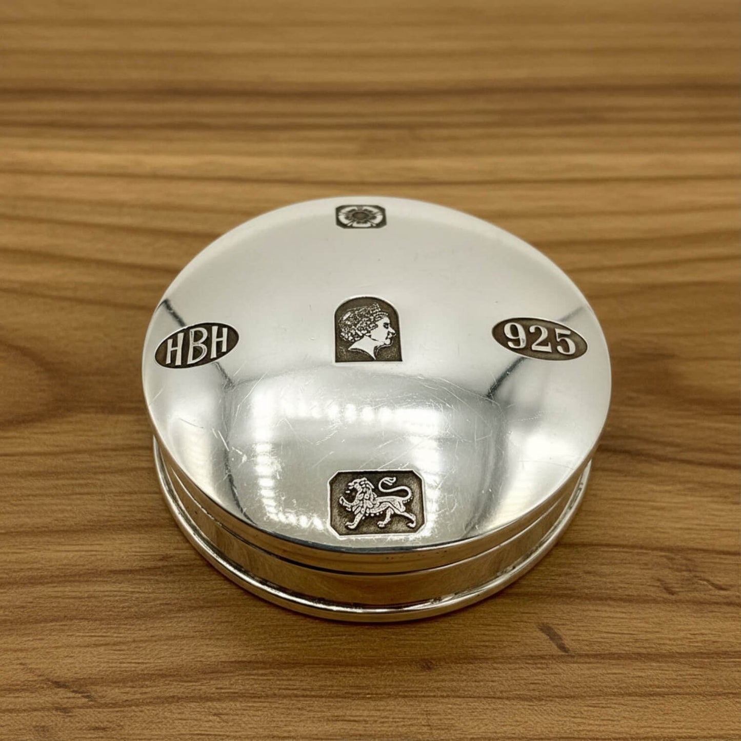 A round sterling silver keepsake box with engraved hallmarks including 'HBH', '925', and lion passant and Queen Elizabeth II motifs, resting on a wooden surface.