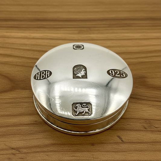 A round sterling silver keepsake box with engraved hallmarks including 'HBH', '925', and lion passant and Queen Elizabeth II motifs, resting on a wooden surface.