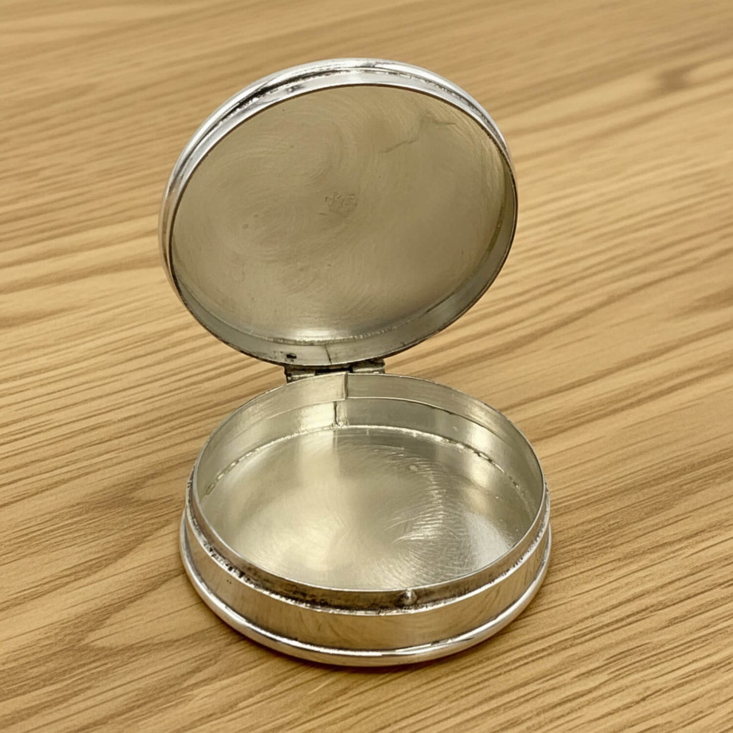 A small round silver  pill box with its lid open, resting on a light brown wooden surface.
