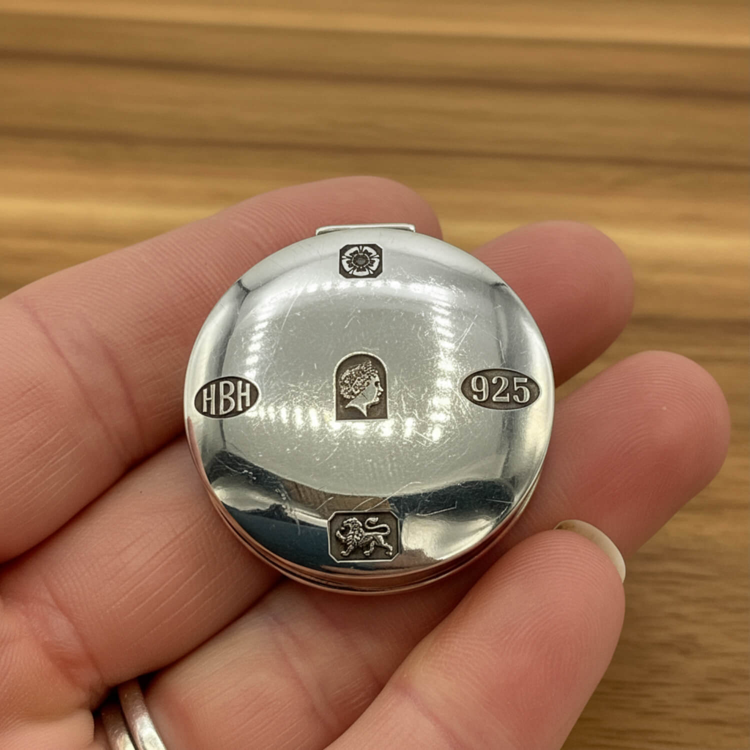 A close-up of a hand holding a round silver pill box, showing sterling silver hallmarks including '925', 'HBH', and Queen Elizabeth's profile.