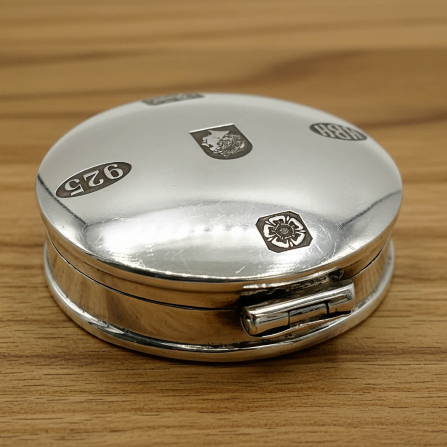 Back of a round sterling silver pill box with engraved hallmarks and a visible hinge, resting on a light brown wooden surface.