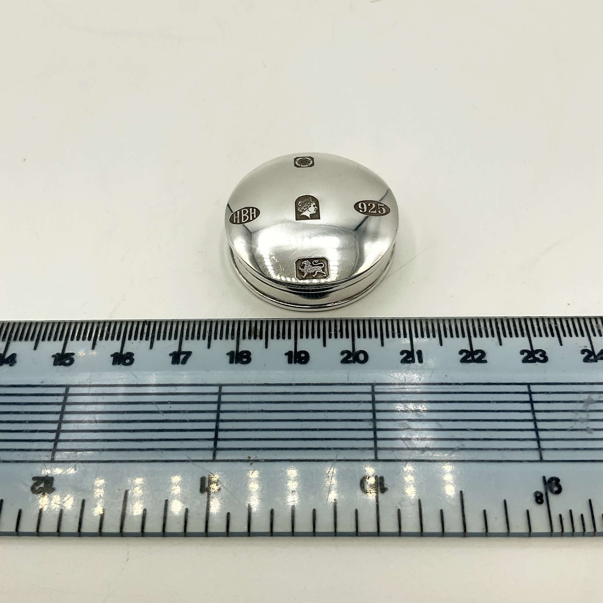 A small round silverpill box with engraved markings, shown above a clear plastic ruler on a white background to indicate size.