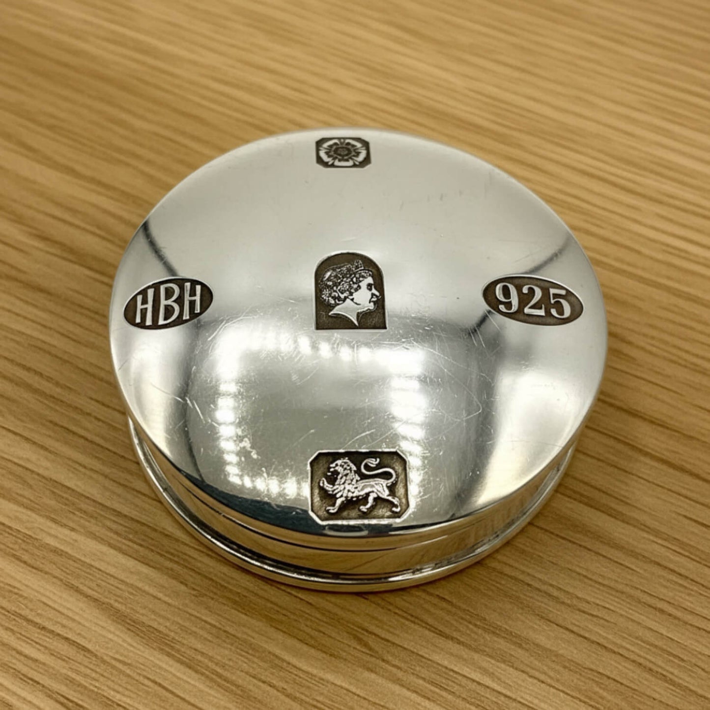A round silverpill box lid with engraved details, including the profile of Queen Elizabeth II, a lion emblem, 'HBH', and '925', resting on a light wood grain surface.