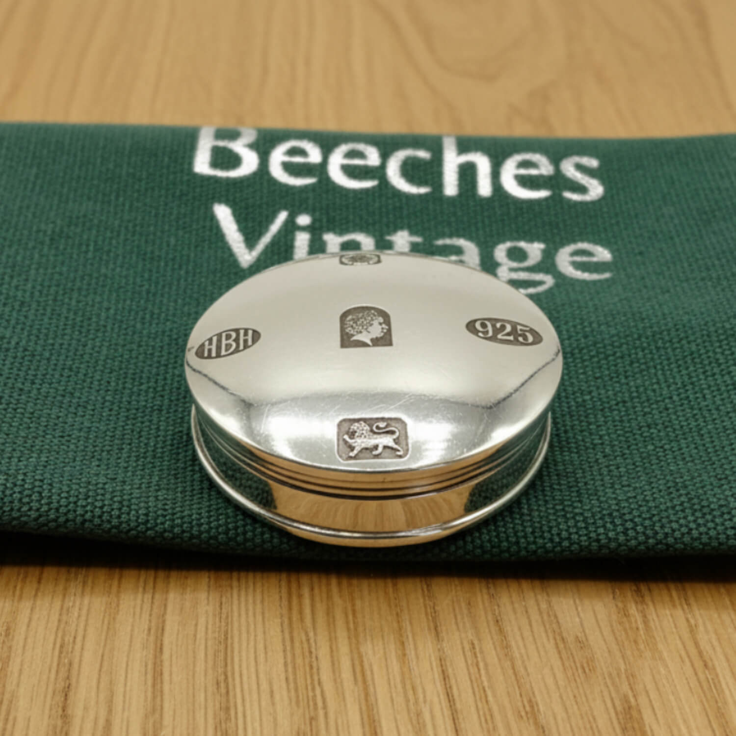 A round silver pill box with engraved hallmarks, resting on a dark green fabric pouch printed with 'Beeches Vintage' on a light wood surface.