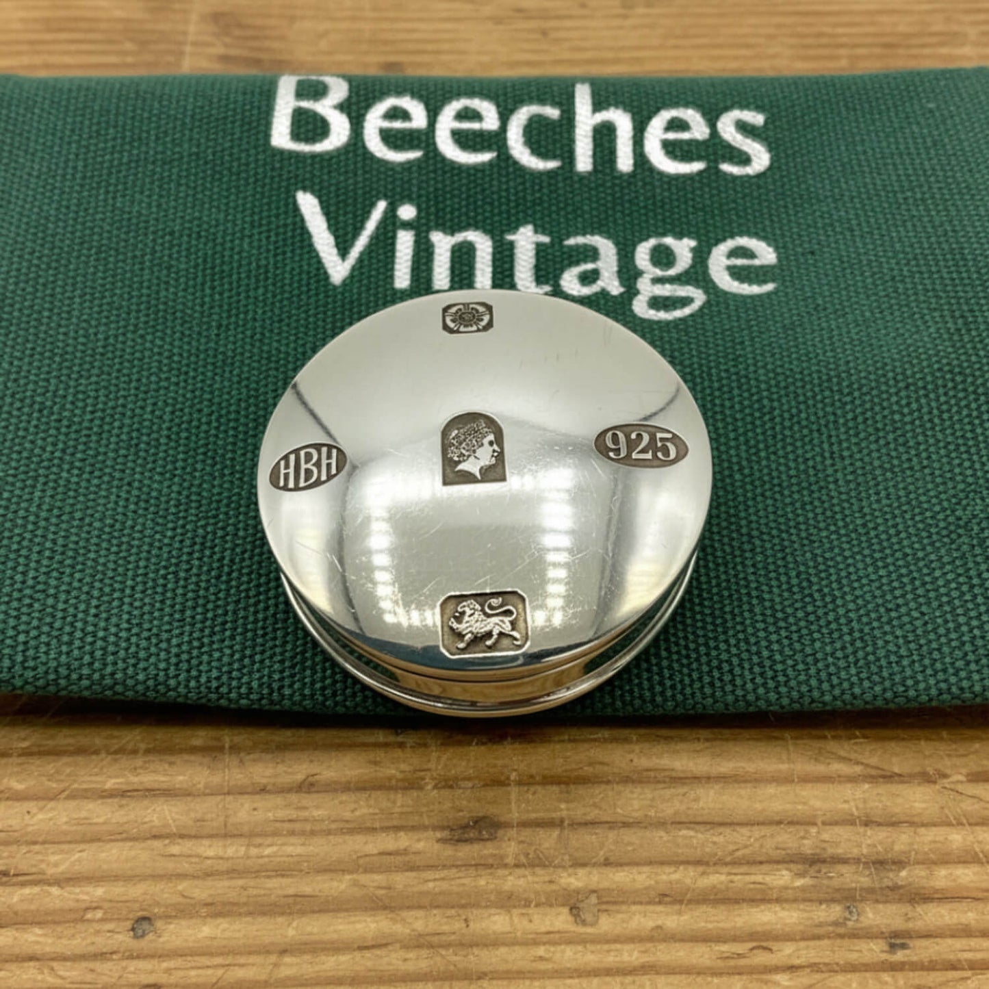 A round silver pill box lid with engraved details, displayed on a green canvas pouch printed with 'Beeches Vintage' on a wooden surface.