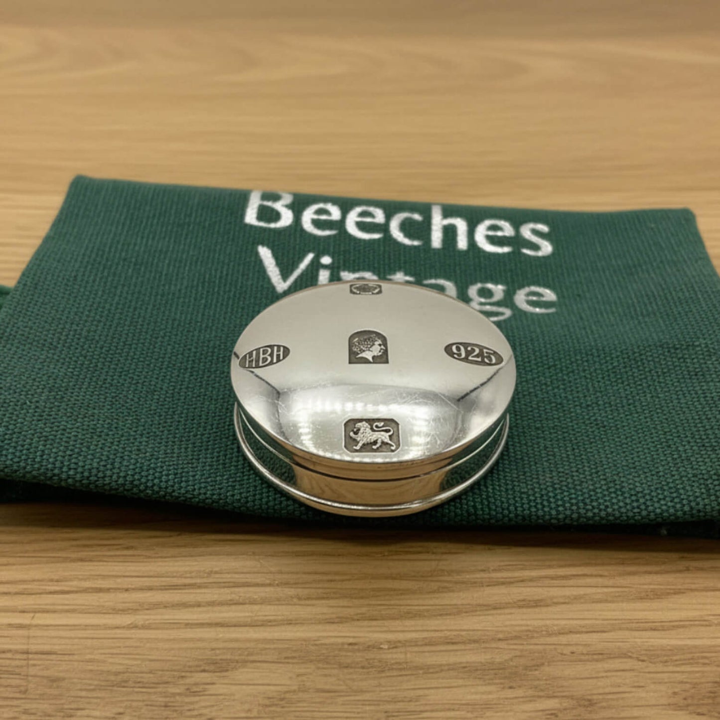 A round silver pill box with engraved hallmarks, resting on a dark green fabric pouch printed with 'Beeches Vintage' on a light wood surface.
