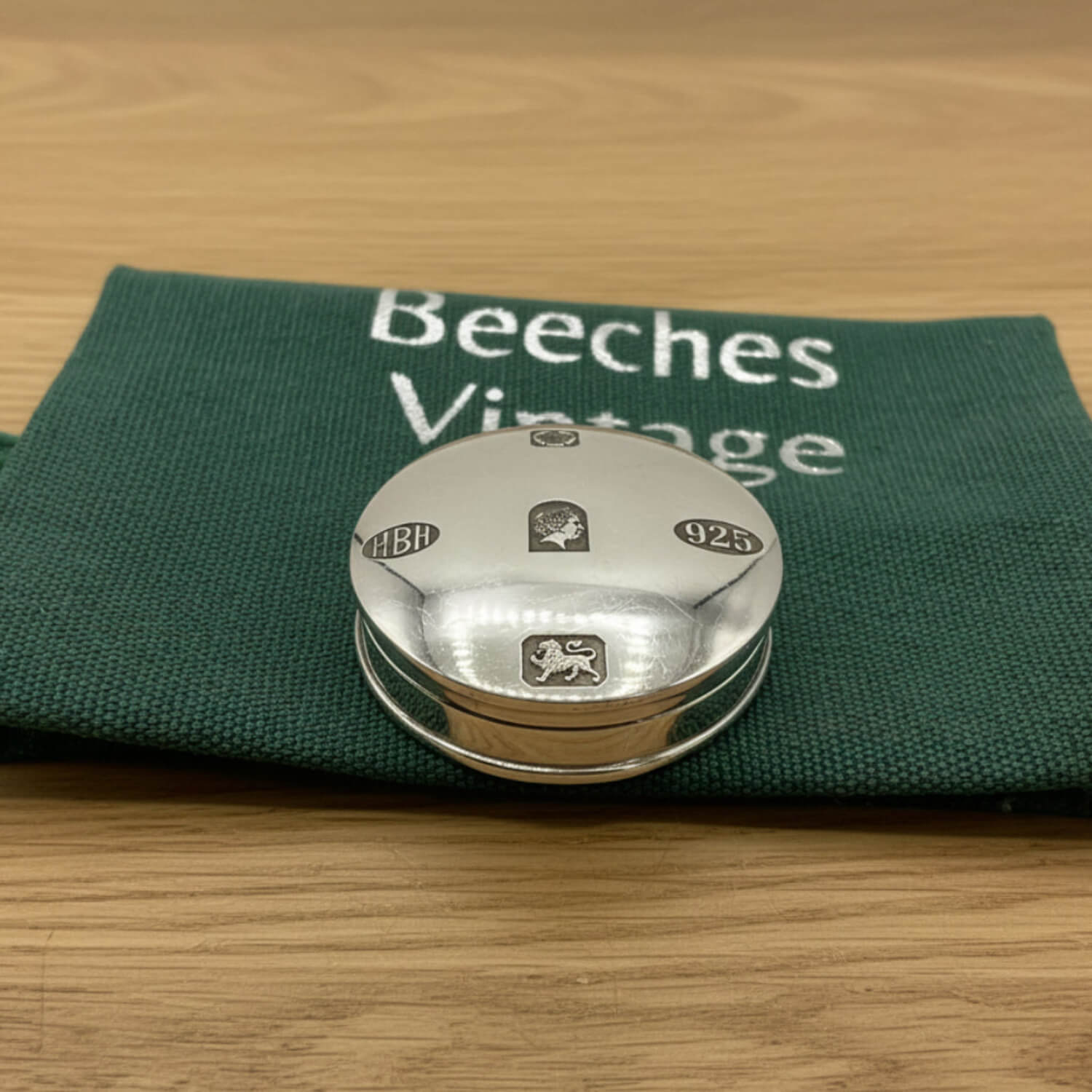 A round silver pill box with engraved hallmarks, resting on a dark green fabric pouch printed with 'Beeches Vintage' on a light wood surface.