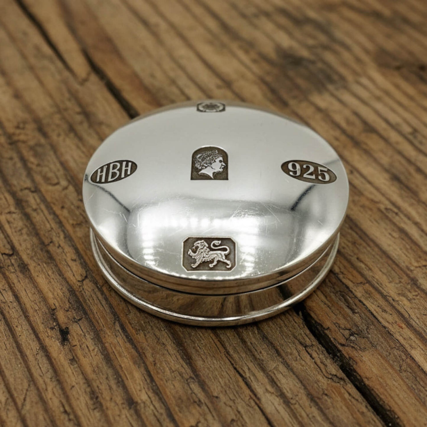 A round sterling silver pill box with engraved details, including the letters 'HBH', a lion passant, and '925', resting on a rustic wooden surface.