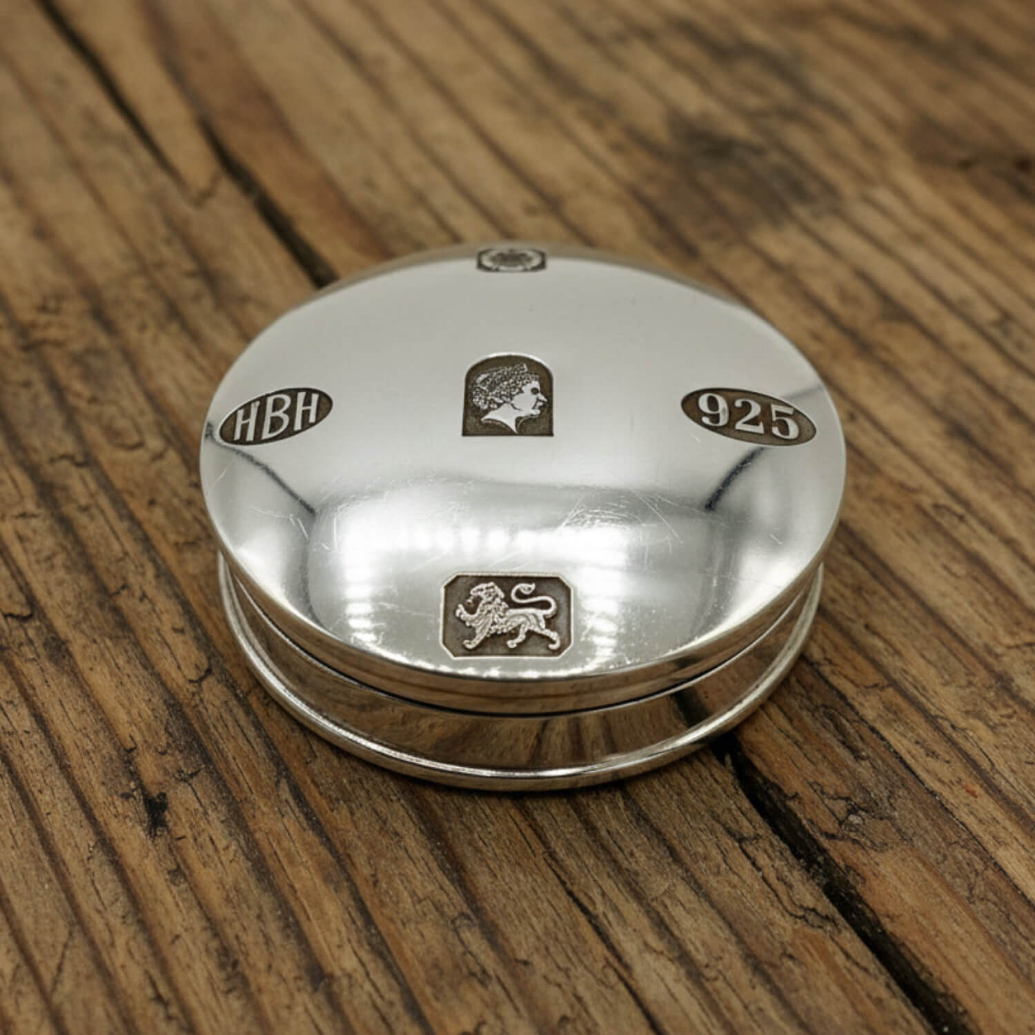 A round sterling silver pill box with engraved details, including the letters 'HBH', a lion passant, and '925', resting on a rustic wooden surface.