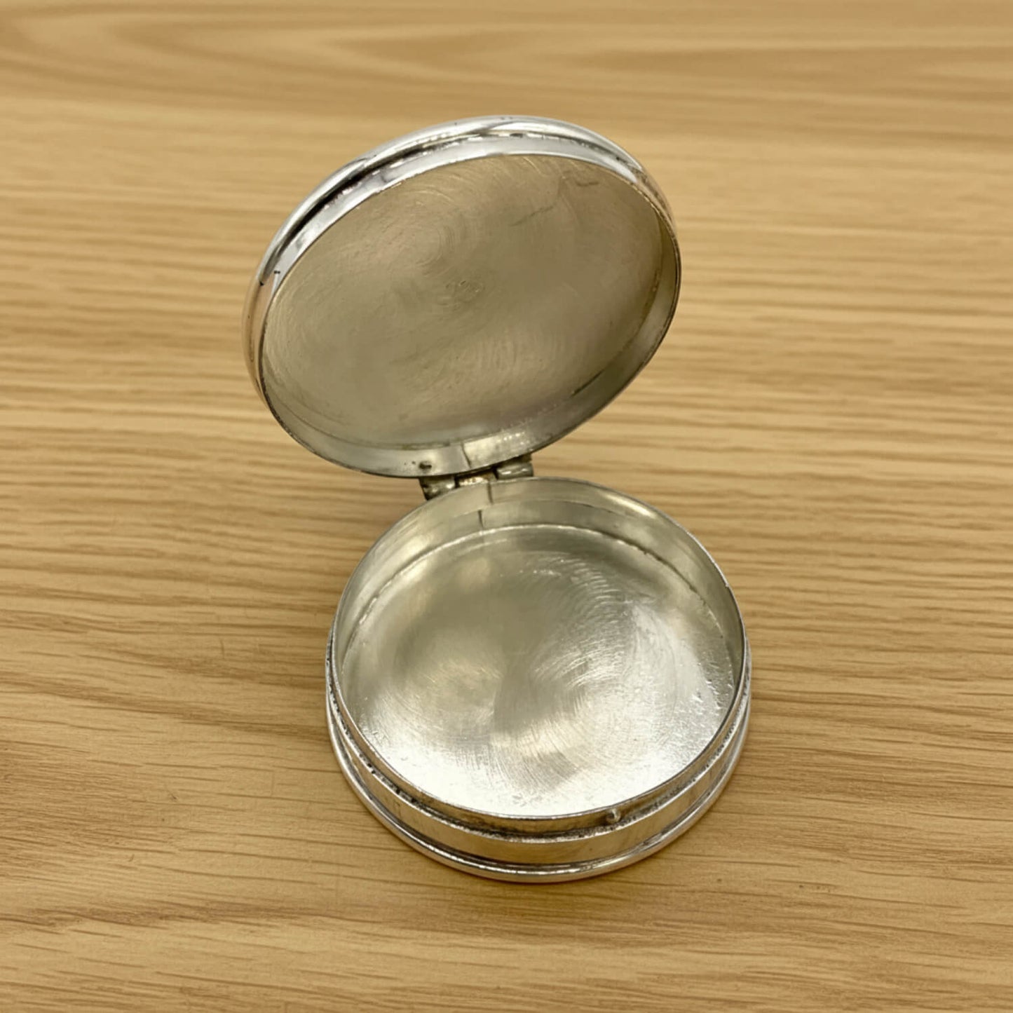 A small round silver pill box with its lid open, resting on a light wood grain surface.