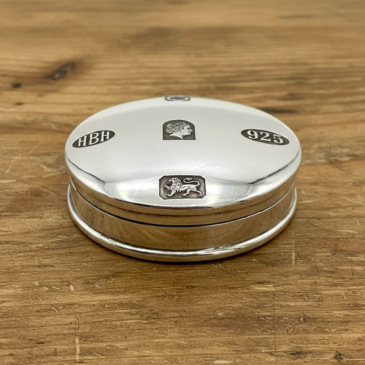 A round sterling silver pill box with engraved details, including 'HBH', '925', and a lion passant, resting on a wooden surface.