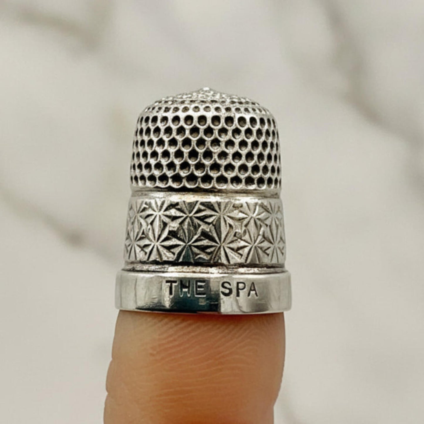 Silver thimble with intricate patterns and 'The Spa' engraving on a finger against a neutral background