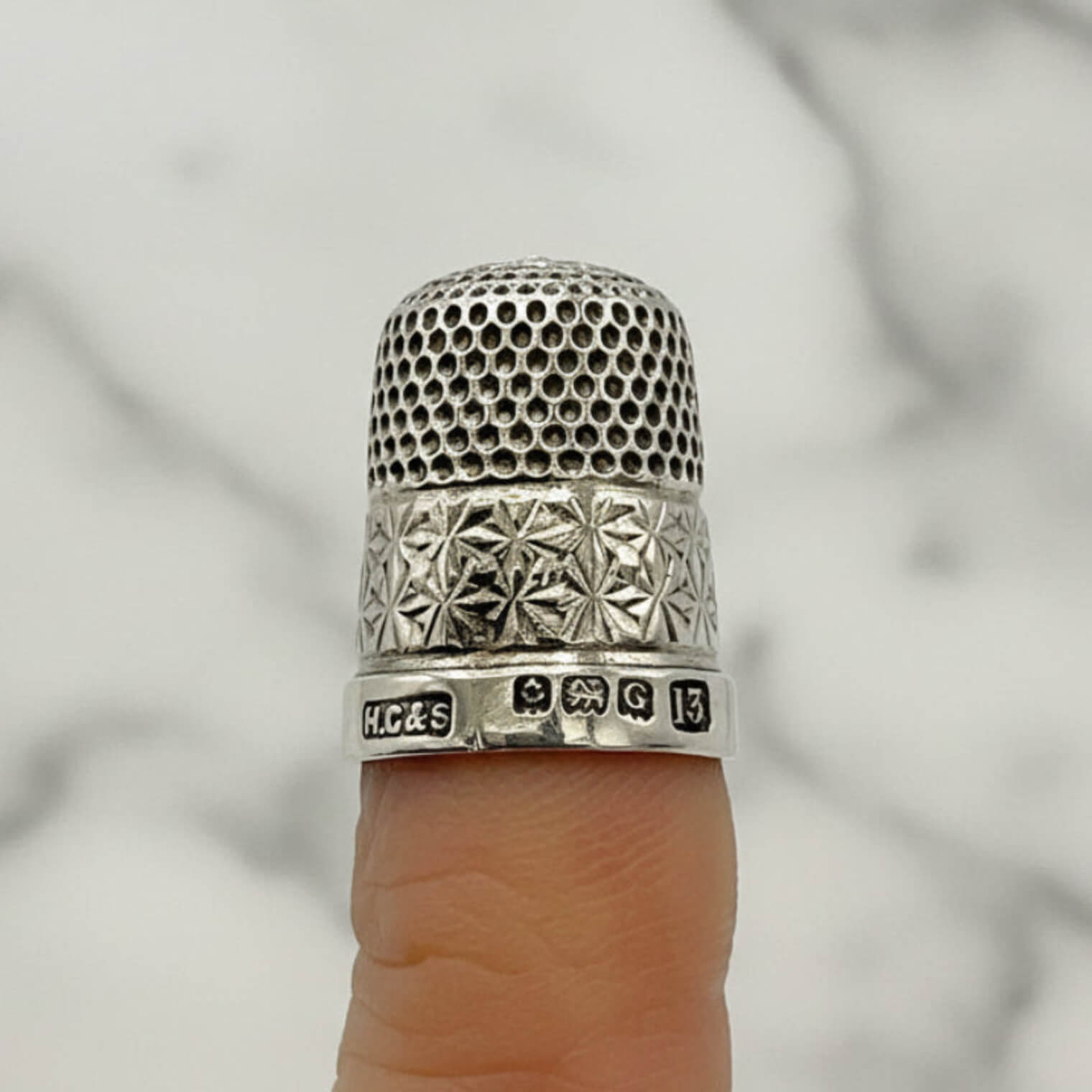 Silver thimble with intricate design on a finger against a neutral background