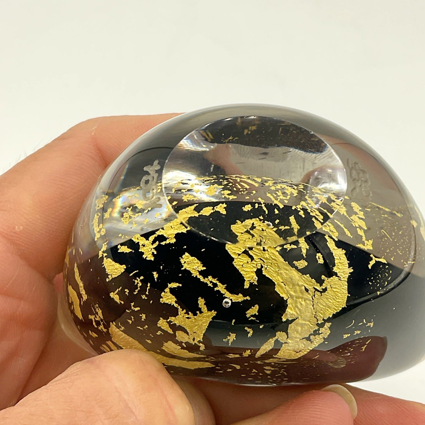 A hand holding a small, oval-shaped clear glass paperweight vase filled with black and gold leaf patterns, set against a plain white background.