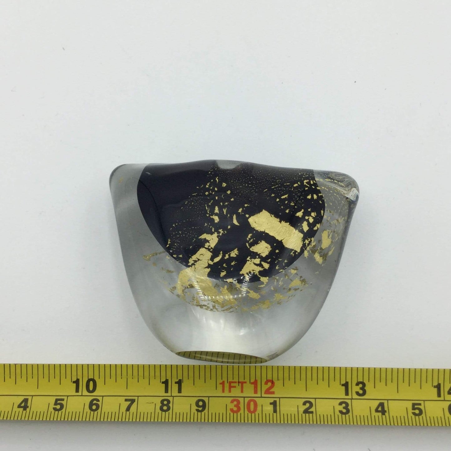 A small organic-shaped clear glass vase with a large black center and scattered gold leaf inclusions, photographed on a white background with a yellow measuring tape ruler along the bottom edge.