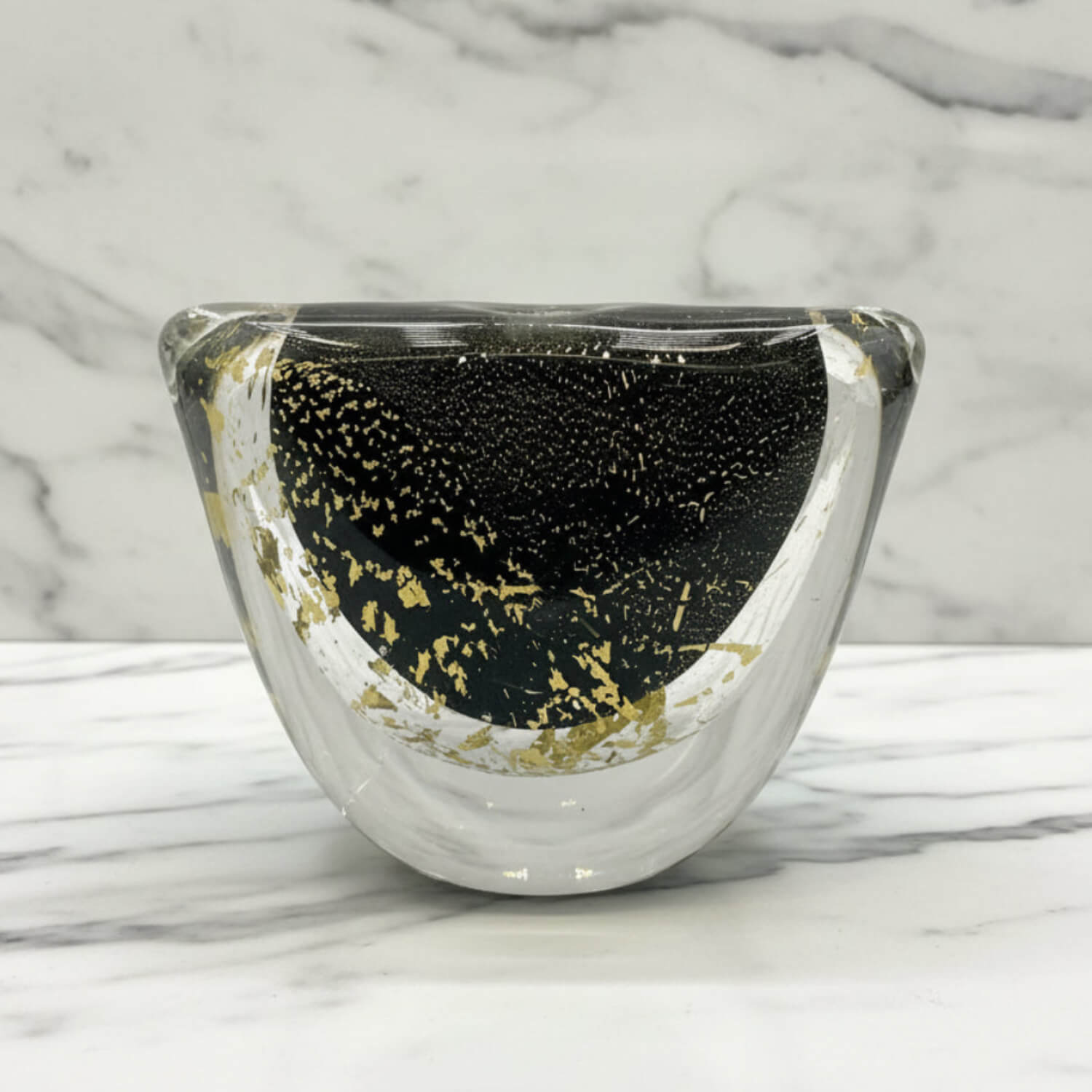 A clear glass vase with a black textured interior and scattered gold leaf flakes, set on a white marble surface against a marble background.