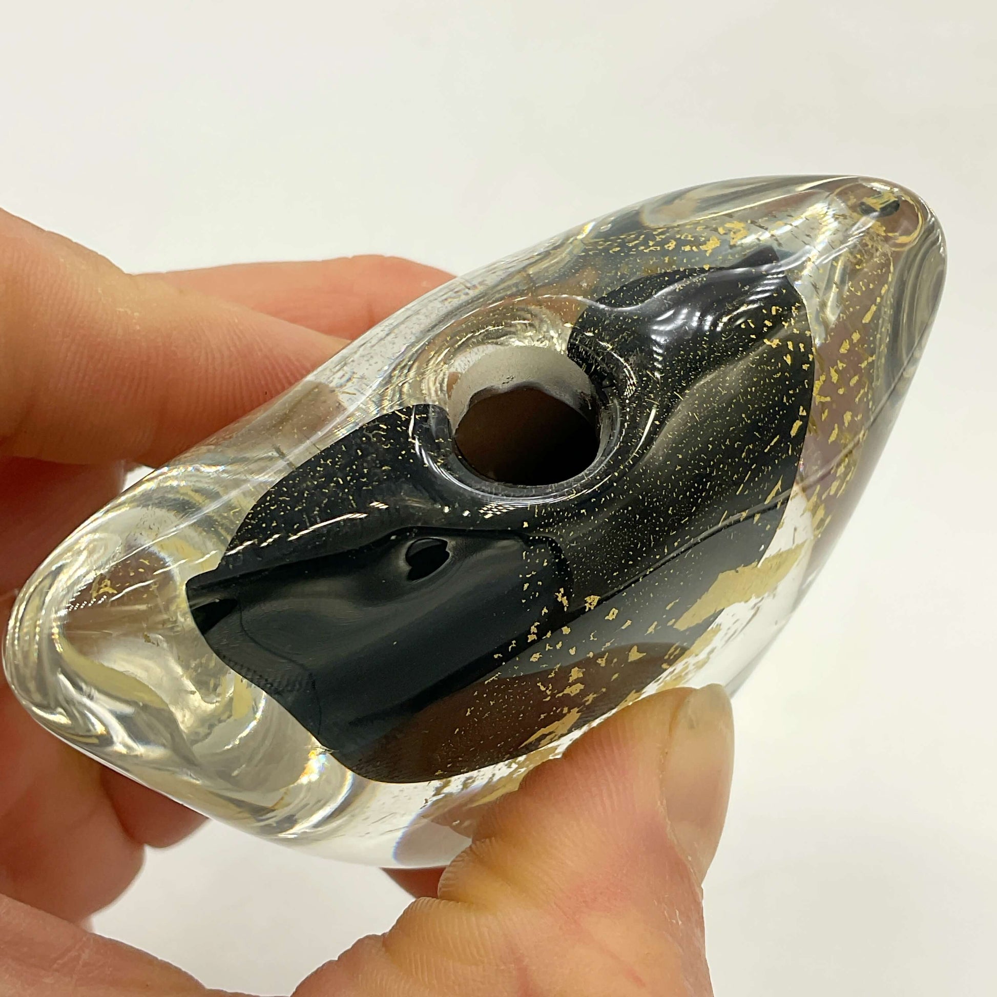 A hand holding a transparent glass vase with a black interior and scattered gold leaf flakes, set against a plain white background.