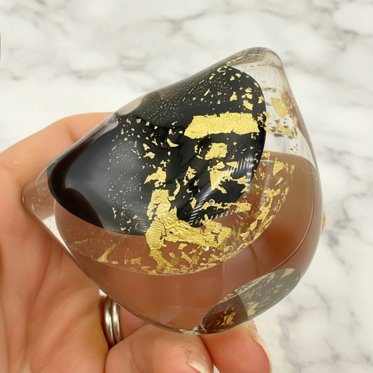 A hand holding a teardrop-shaped clear glass art vase with a glossy black top section, a warm brown lower section, and scattered metallic gold leaf details, set against a light marble background.