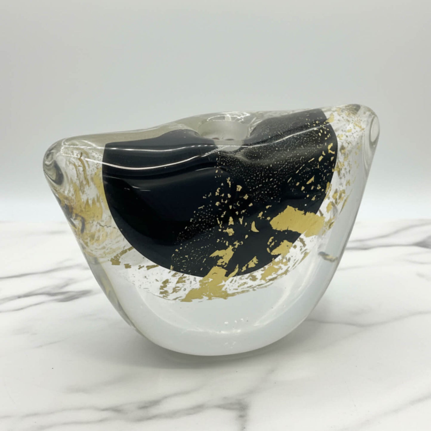 A modern abstract glass vase with a wavy, organic shape, featuring a prominent glossy black central section, gold foil accents, and a clear outer glass body, displayed on a white marble surface against a light gray background.