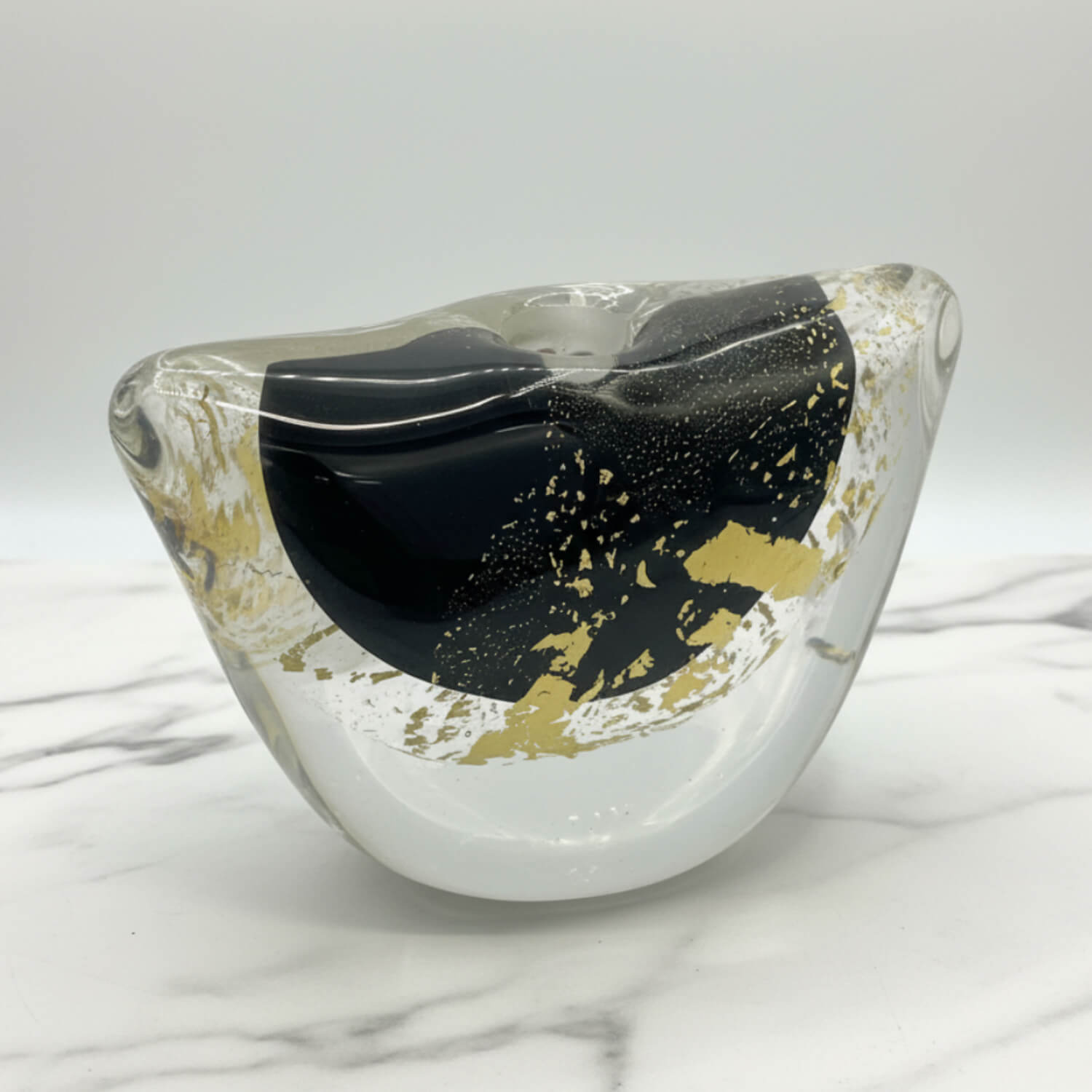 A modern abstract glass vase with a wavy, organic shape, featuring a prominent glossy black central section, gold foil accents, and a clear outer glass body, displayed on a white marble surface against a light gray background.