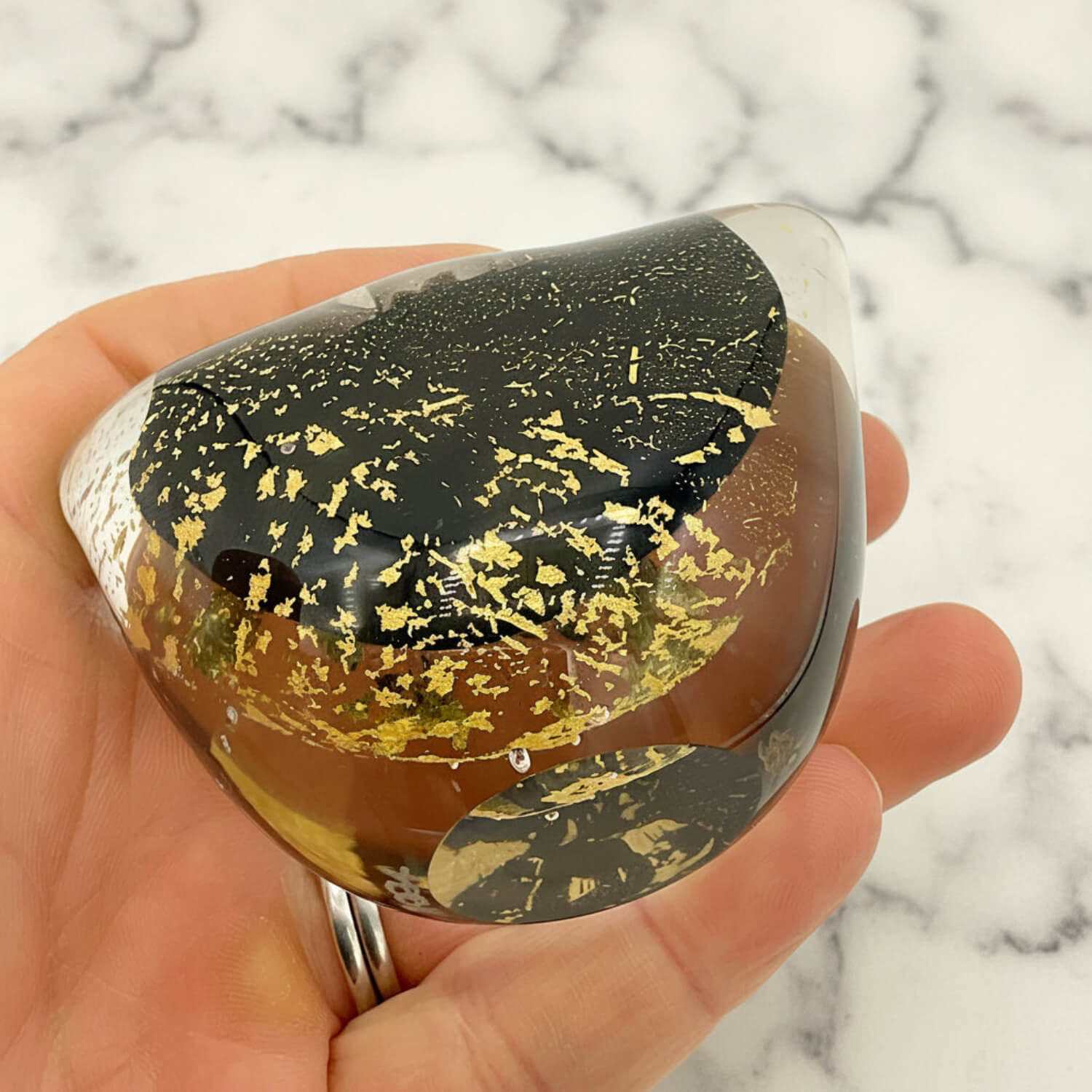 A hand holds a teardrop-shaped clear resin lamp or decorative vase featuring a black top half with gold leaf flecks, a brown middle band, and a silver metallic edge, set against a white marble background.