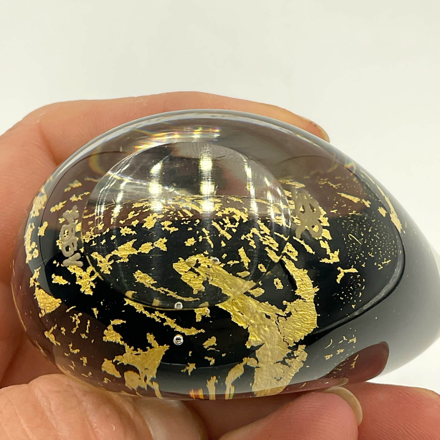 A hand holds a glossy, oval-shaped black resin or glass-like vase decorated with scattered metallic gold leaf flakes, photographed against a plain light background.