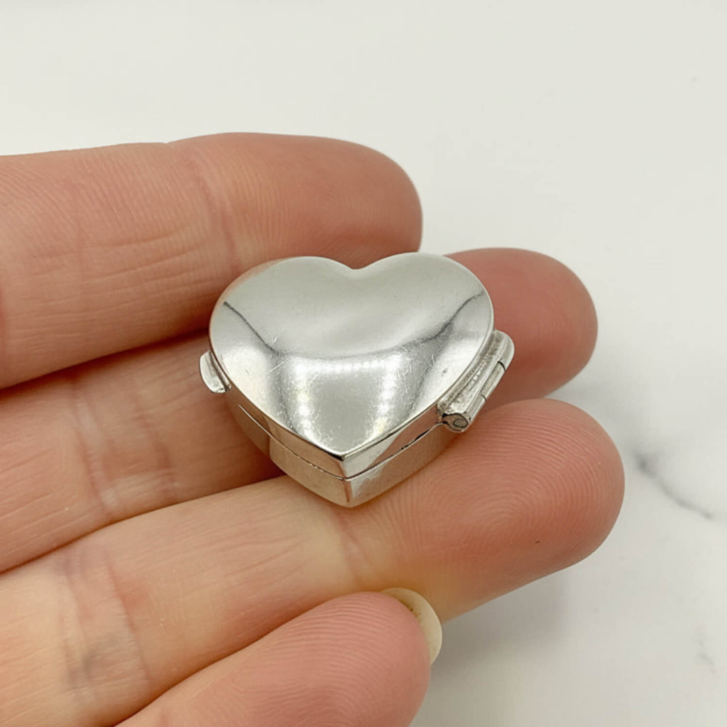 Silver heart-shaped pill box held in a hand against a white background