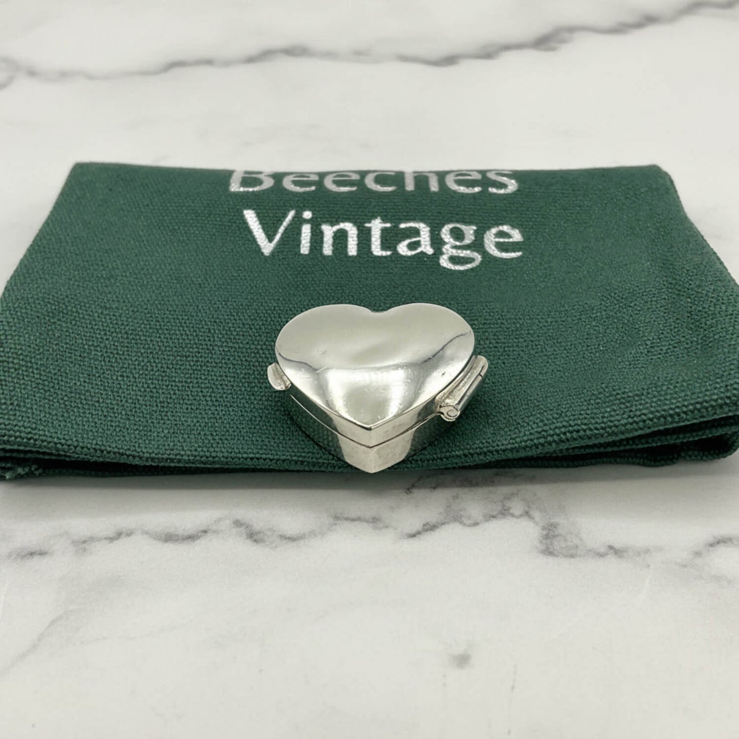 Heart-shaped silver pill box on a green cloth bag with 'Beeches Vintage' branding, on a marble surface.