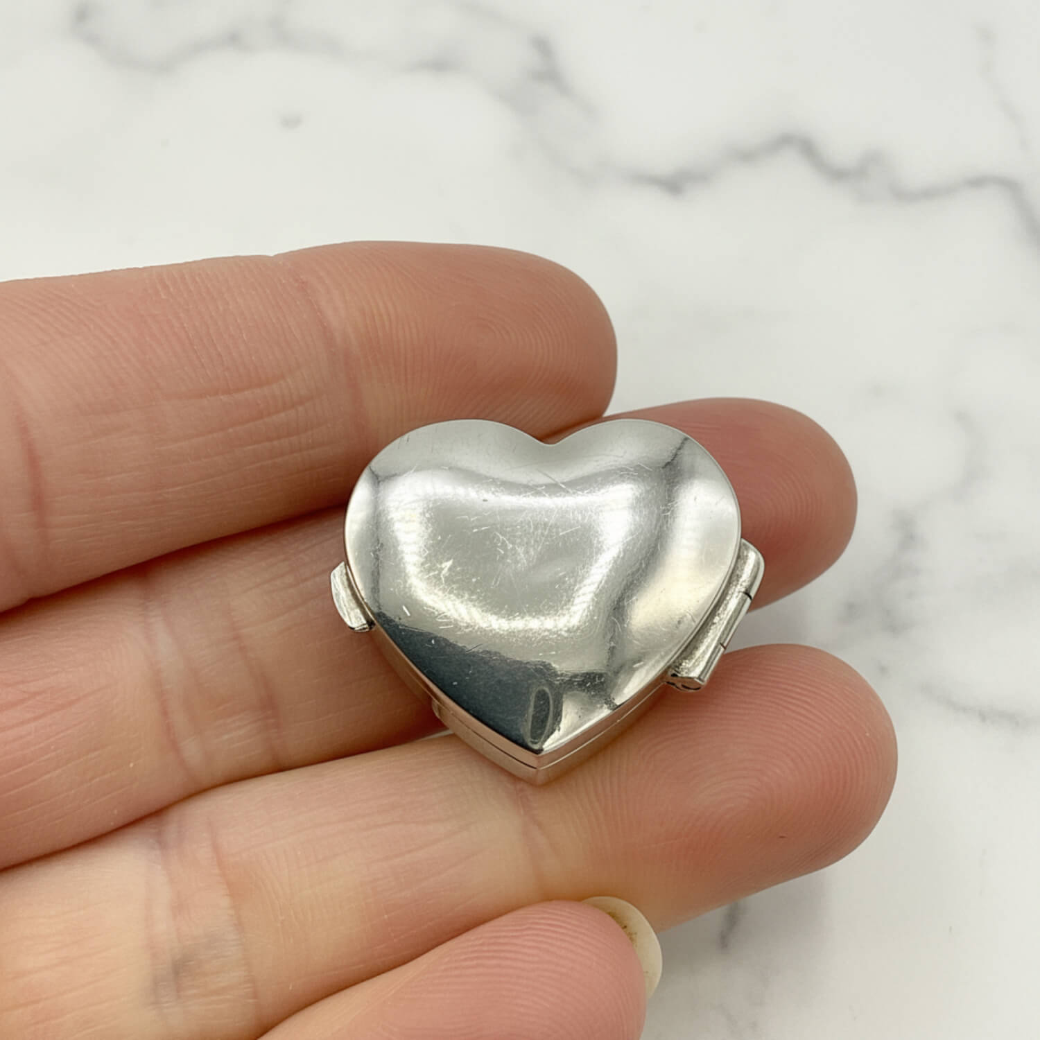 Silver heart-shaped pill box held between fingers on a white background