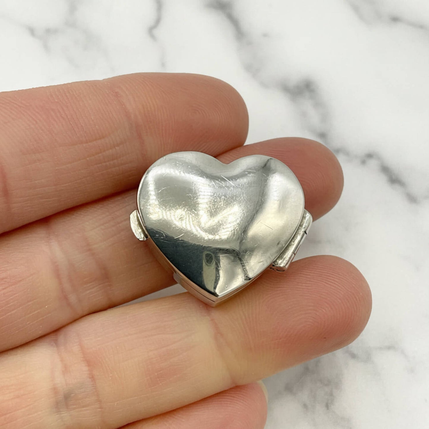 Silver heart-shaped pill box held between fingers on a marble background