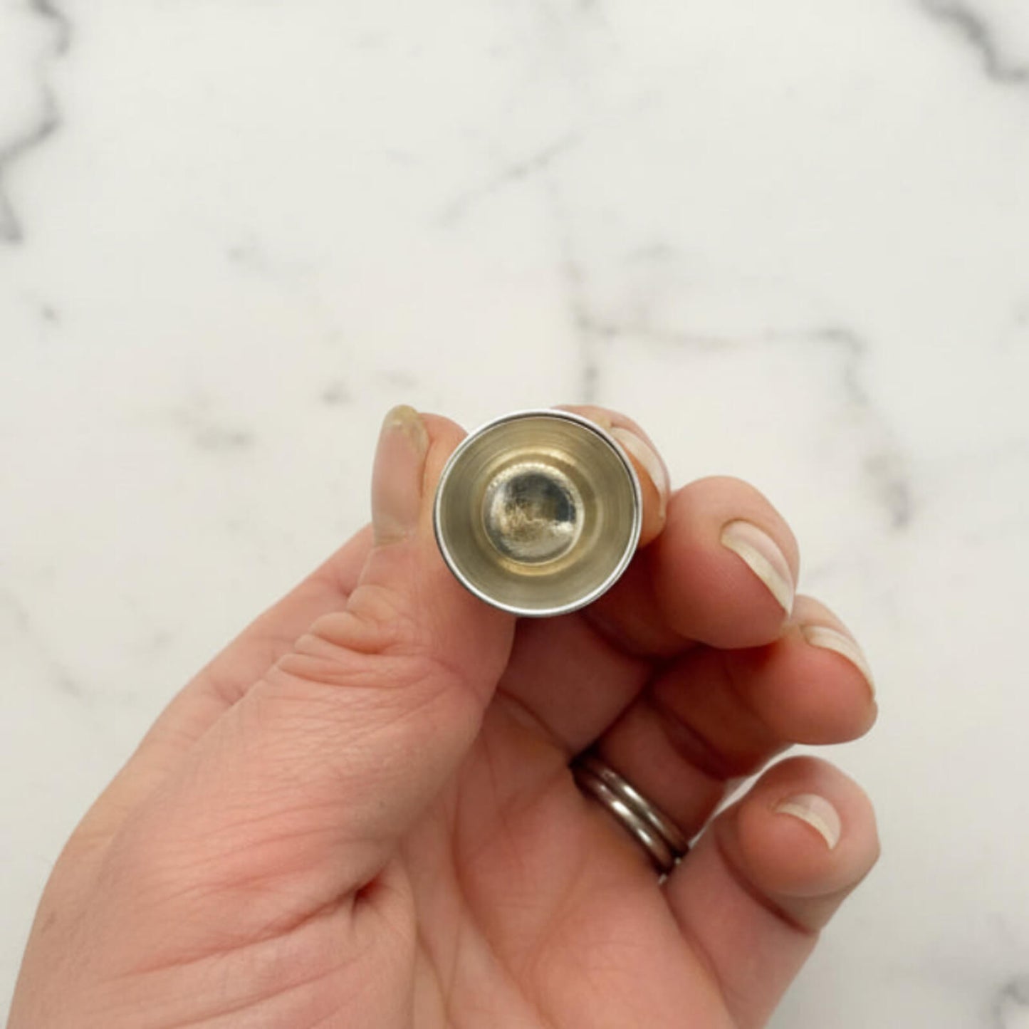 Hand holding a small silver thimble showing the inside, against a marble background