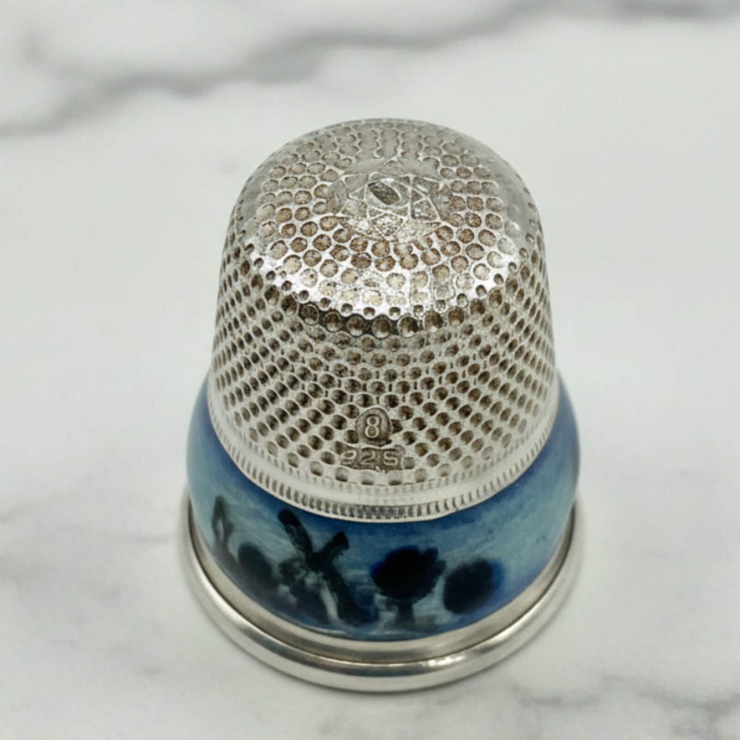 Silver thimble with a blue windmill enamel pattern on a marble surface