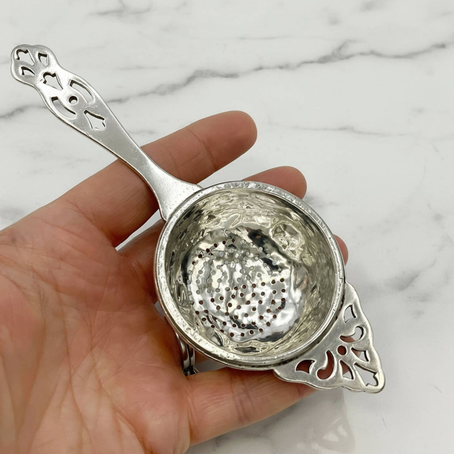 Silver tea strainer held in a hand against a marble background