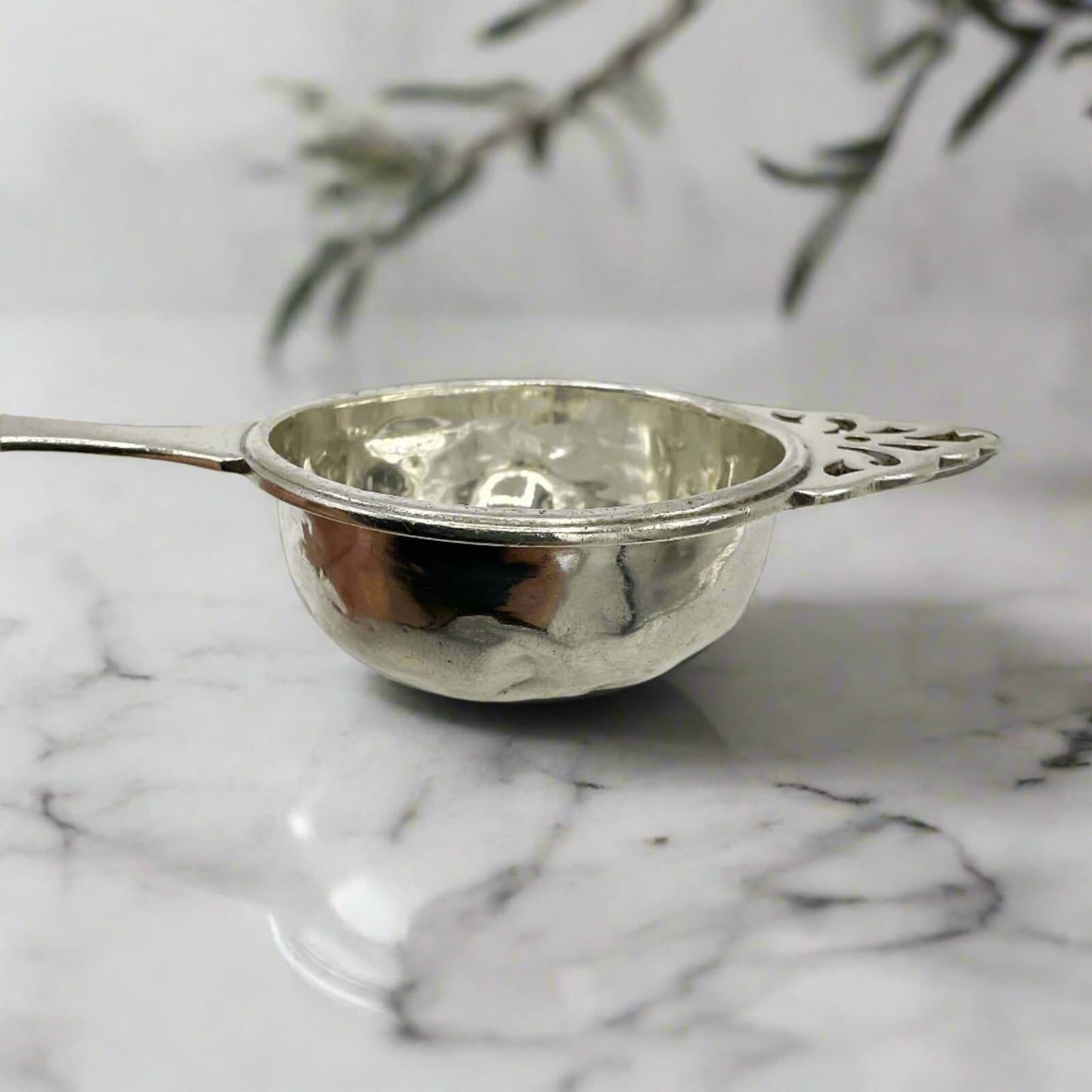 Silver tea strainer on a marble surface with a blurred plant in the background