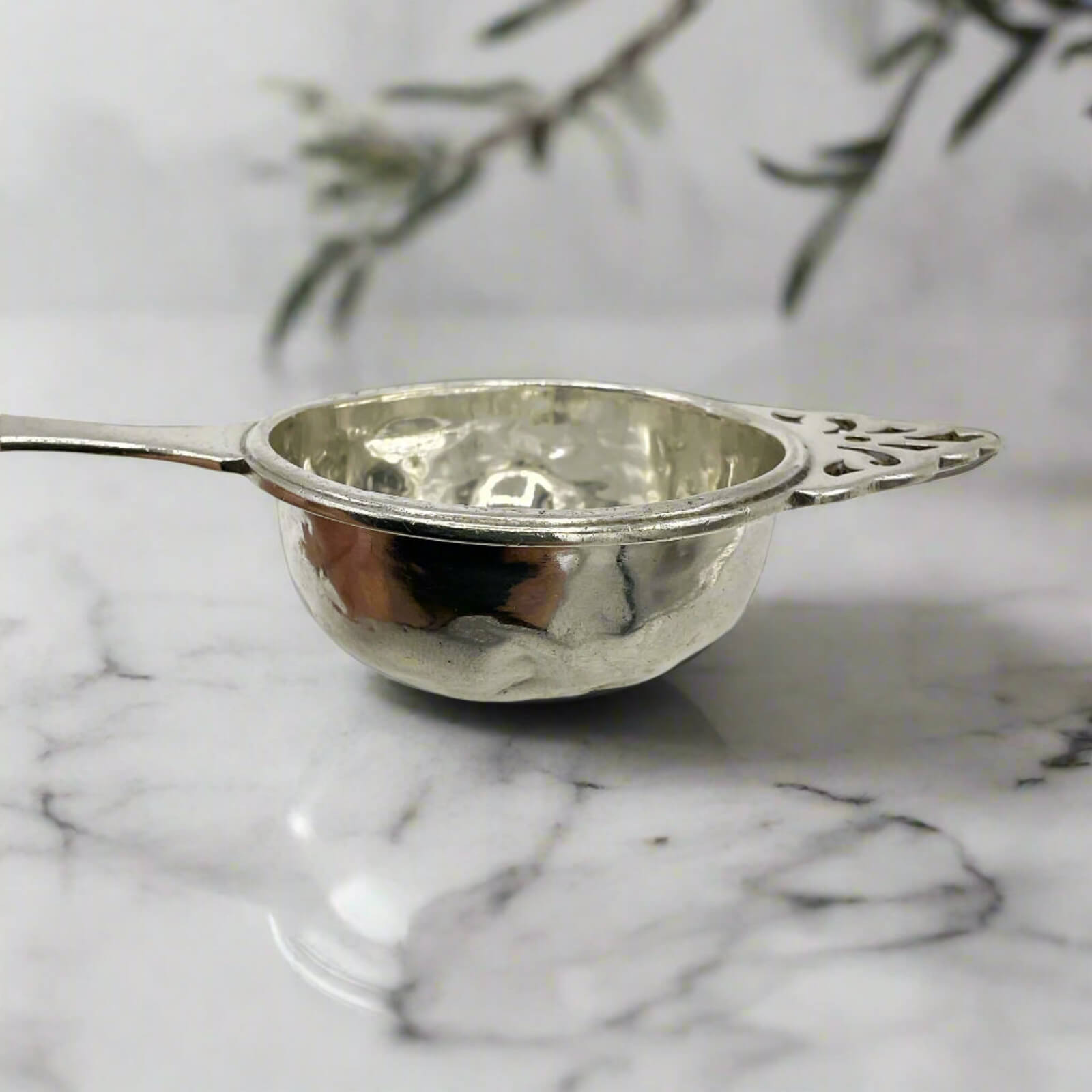 Silver tea strainer on a marble surface with a blurred plant in the background
