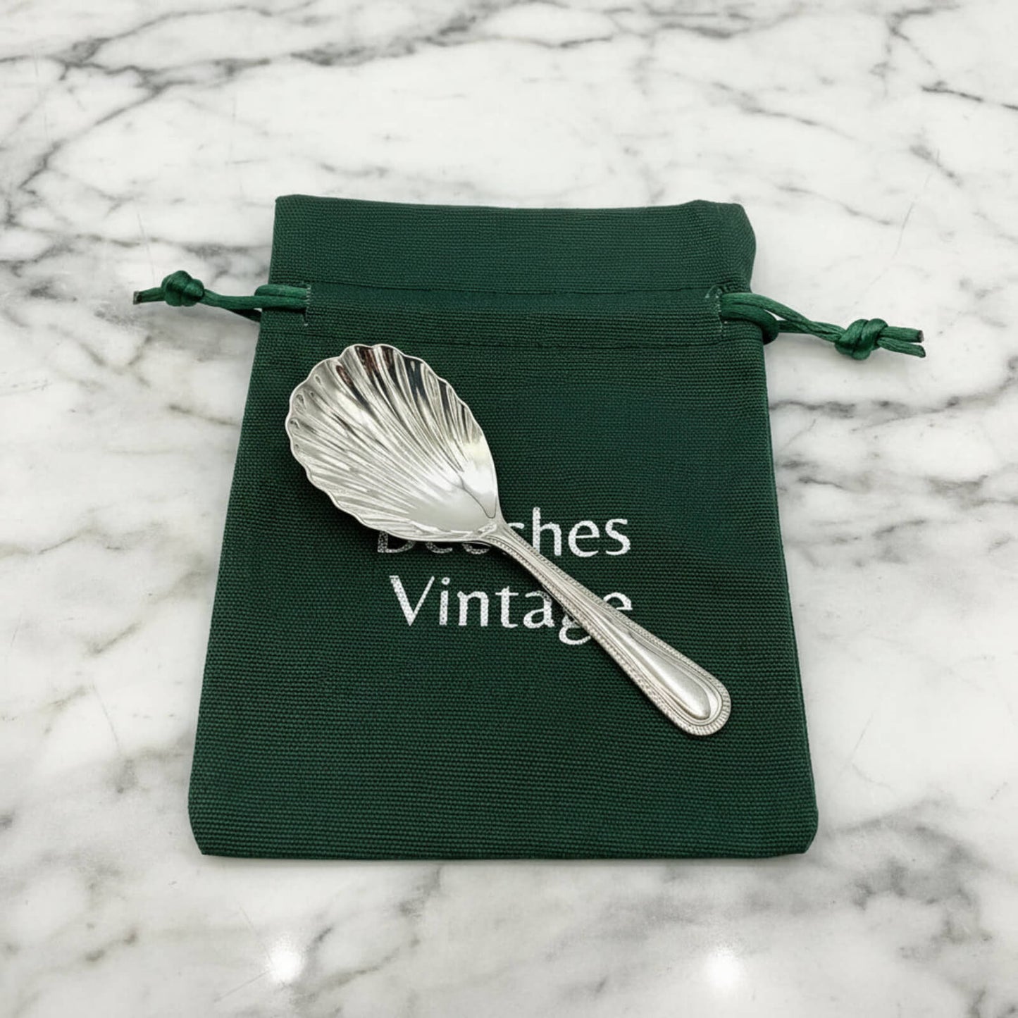 Silver shell-shaped caddy spoon on a green drawstring bag with 'Beeches Vintage' text on a marble background