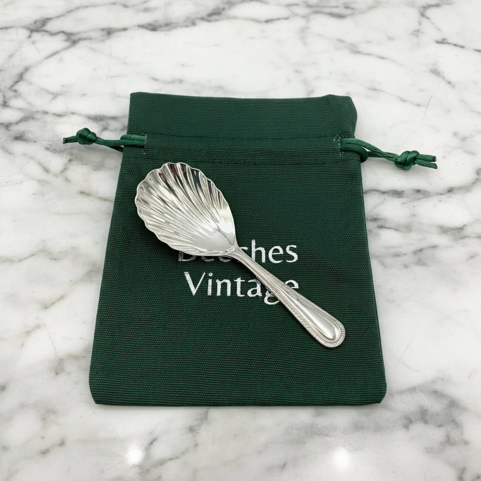 Silver shell-shaped caddy spoon on a green drawstring bag with 'Beeches Vintage' text on a marble background