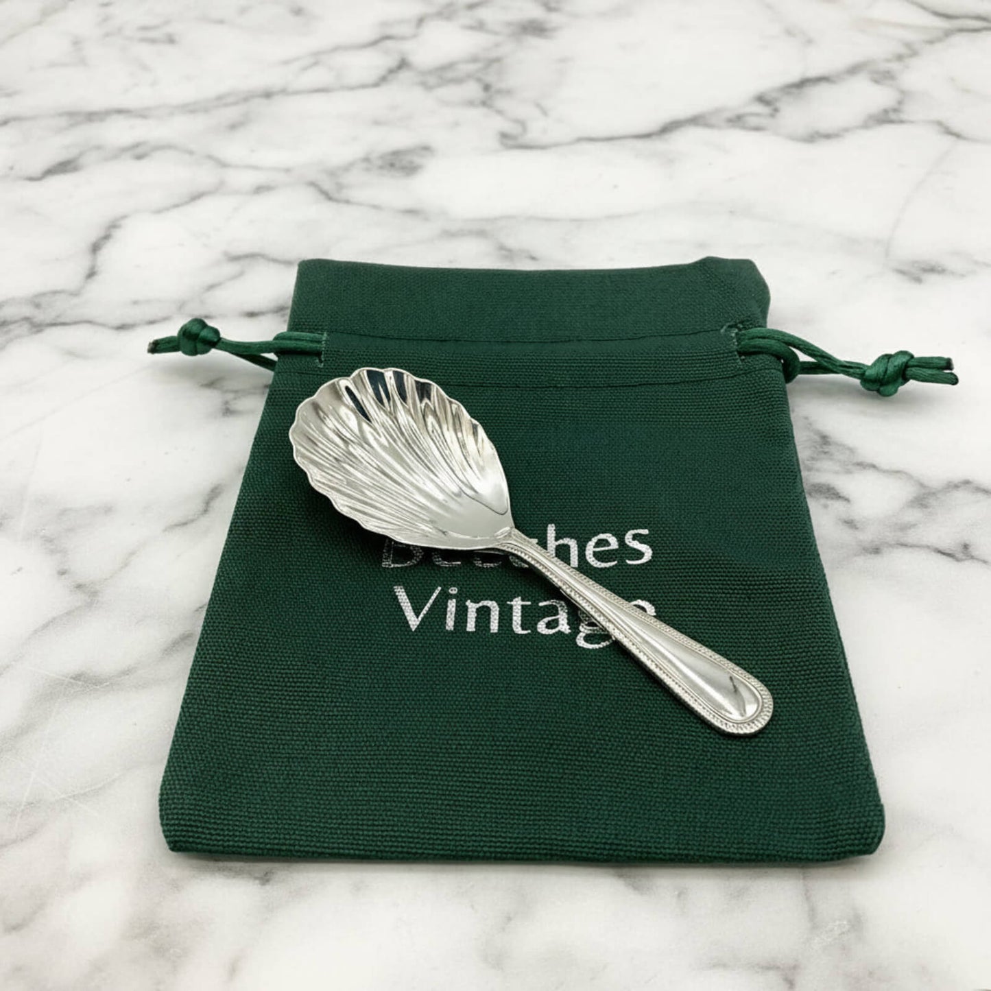 Silver shell-shaped spoon on a green drawstring bag with 'Beeches Vintage' text on a marble background