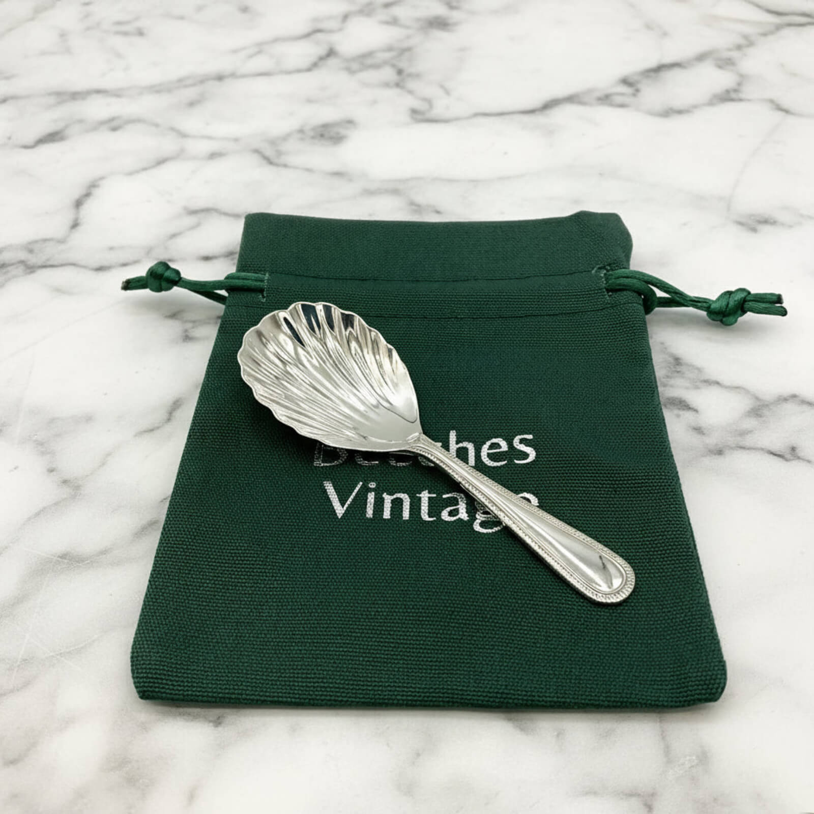 Silver shell-shaped spoon on a green drawstring bag with 'Beeches Vintage' text on a marble background