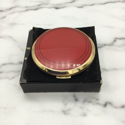 A round red compact case with a gold-tone metal rim, resting on a black velvet-lined box over a white marble surface.