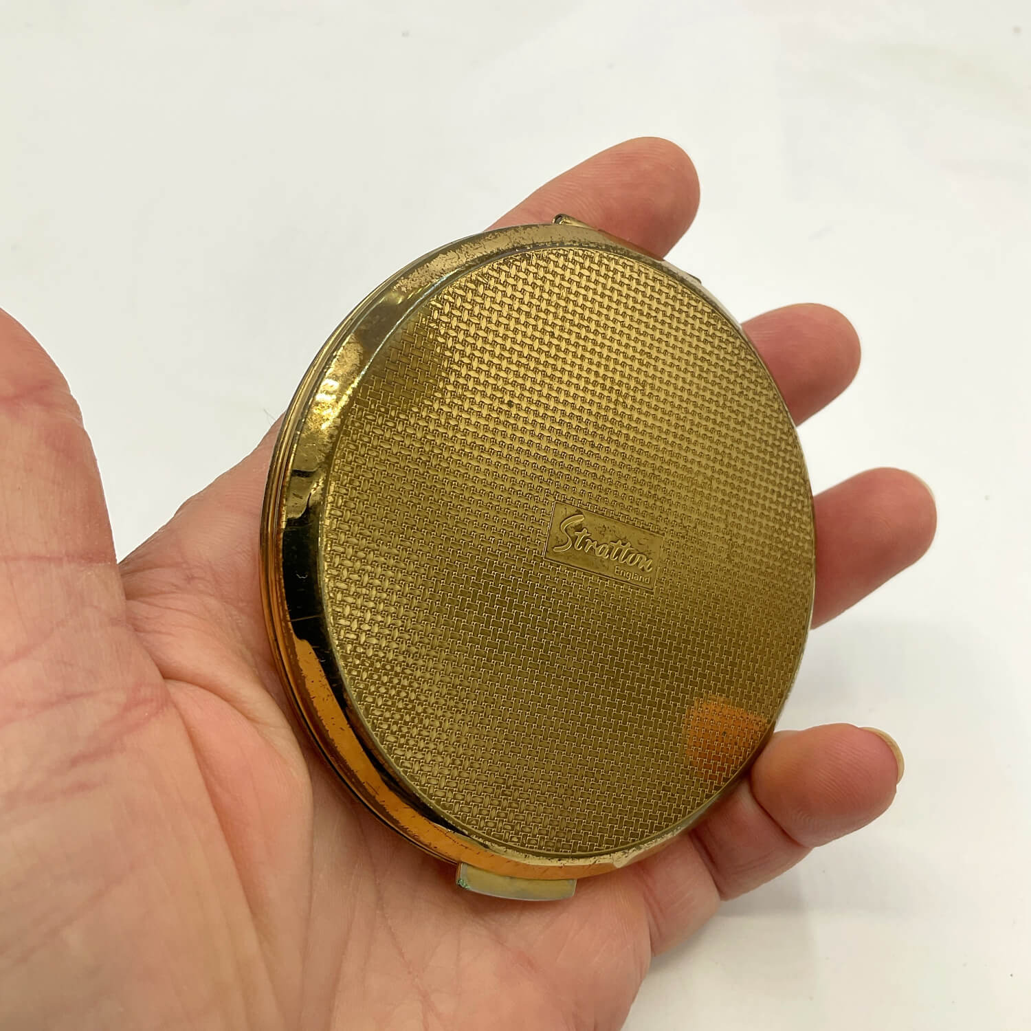 A vintage gold compact mirror with a textured mesh cover and an embossed 'Greeting' logo, held in a person's hand against a plain white background.