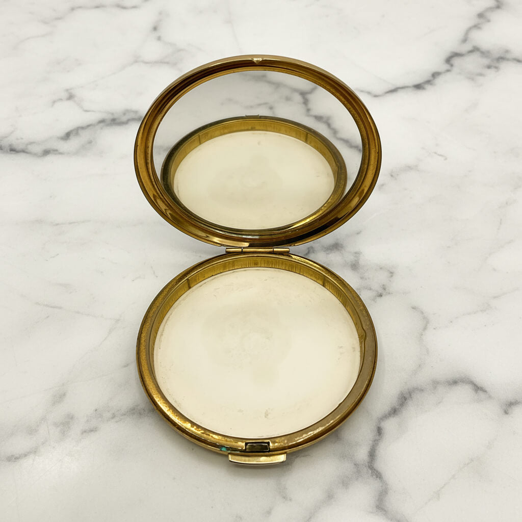 An open round compact case with a polished gold-tone metal frame and mirror lid, resting on a white marble surface with gray veining.