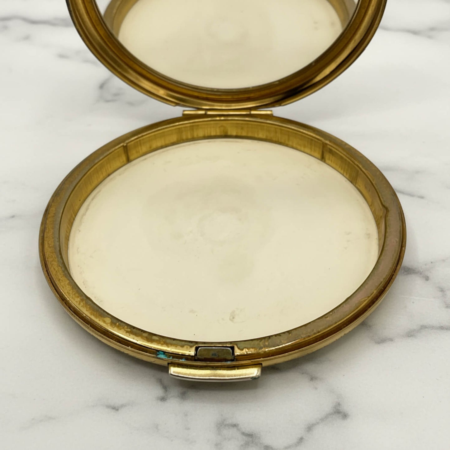 An open round compact with a gold-toned metal frame and mirror lid, showing a light cream-colored pressed powder, set on a white marble surface.